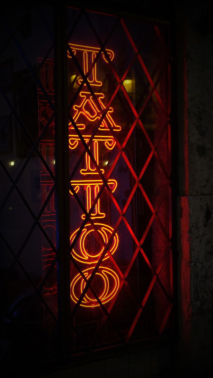 gallery-1 Vibrant neon tattoo sign behind lattice window in Lisbon, Portugal at night.