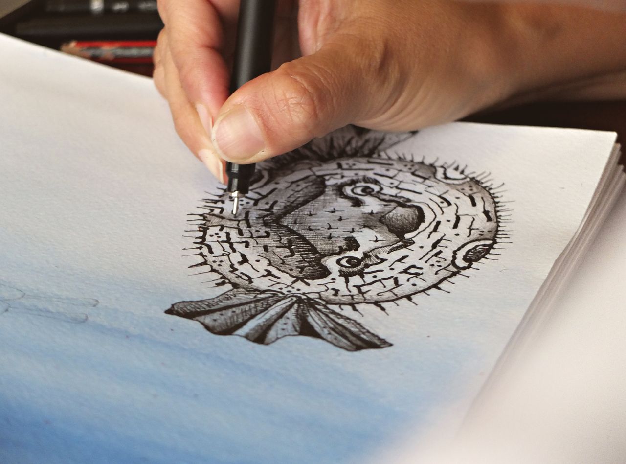 services-01 Close-up of a hand sketching a detailed pufferfish illustration on paper.