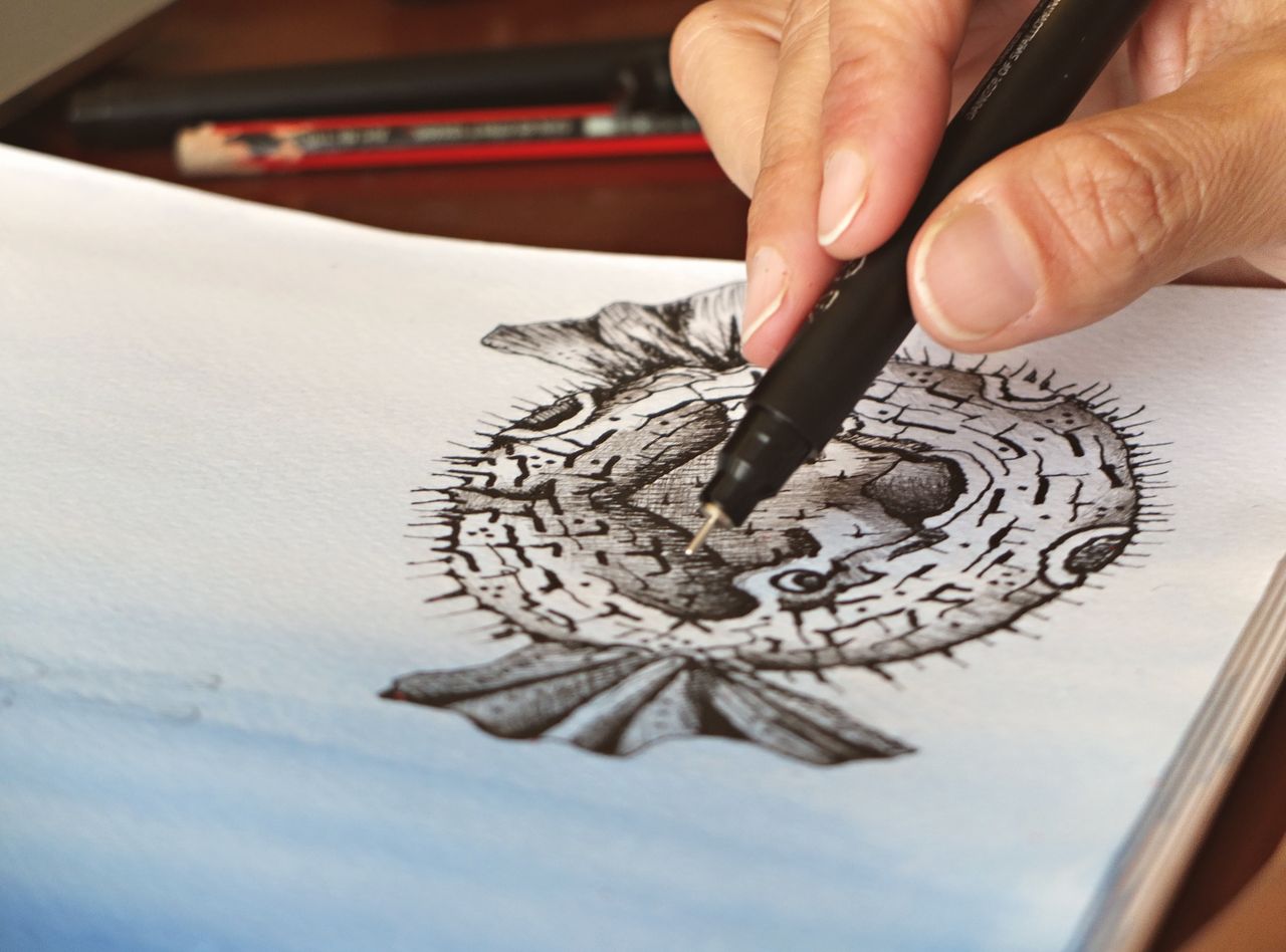 hero-img-01 A person skillfully drawing a pufferfish illustration with fine details using a pen.