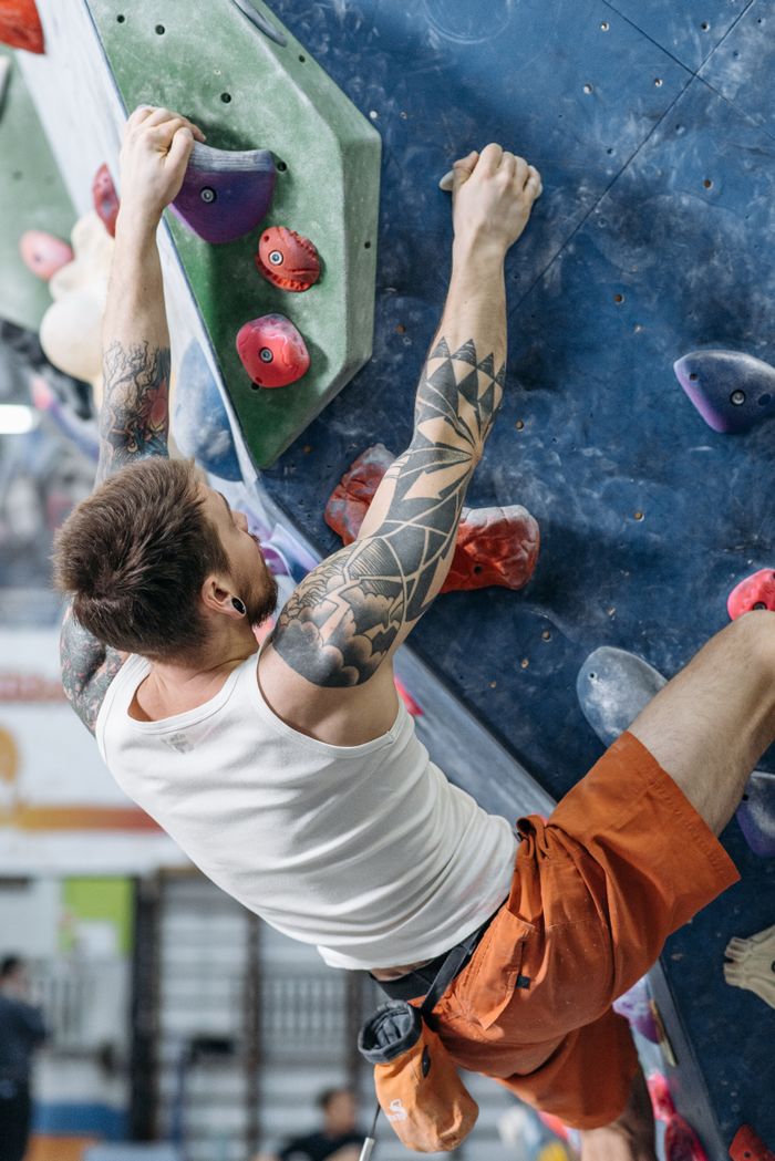 Tattooed man climbing indoor rock wall in orange shorts, focused on grips.
